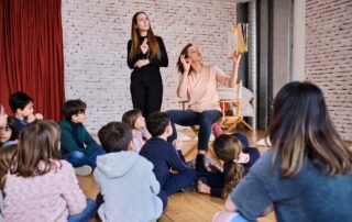 All’interno di una sala con le pareti a mattoncini color beige, un gruppo di bambini seduto sul pavimento sta ascoltando una storia che viene letta da Valeria e tradotta in LIS da Sara