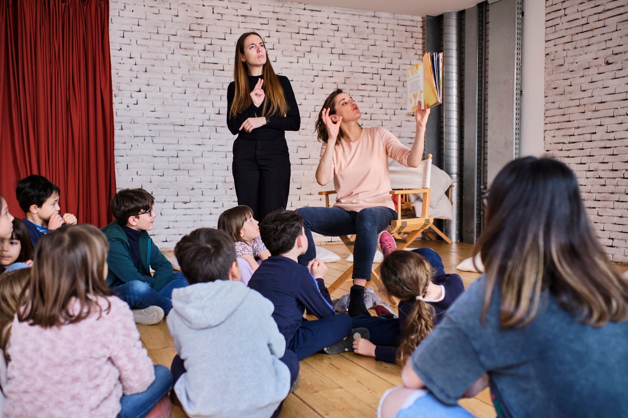 All’interno di una sala con le pareti a mattoncini color beige, un gruppo di bambini seduto sul pavimento sta ascoltando una storia che viene letta da Valeria e tradotta in LIS da Sara