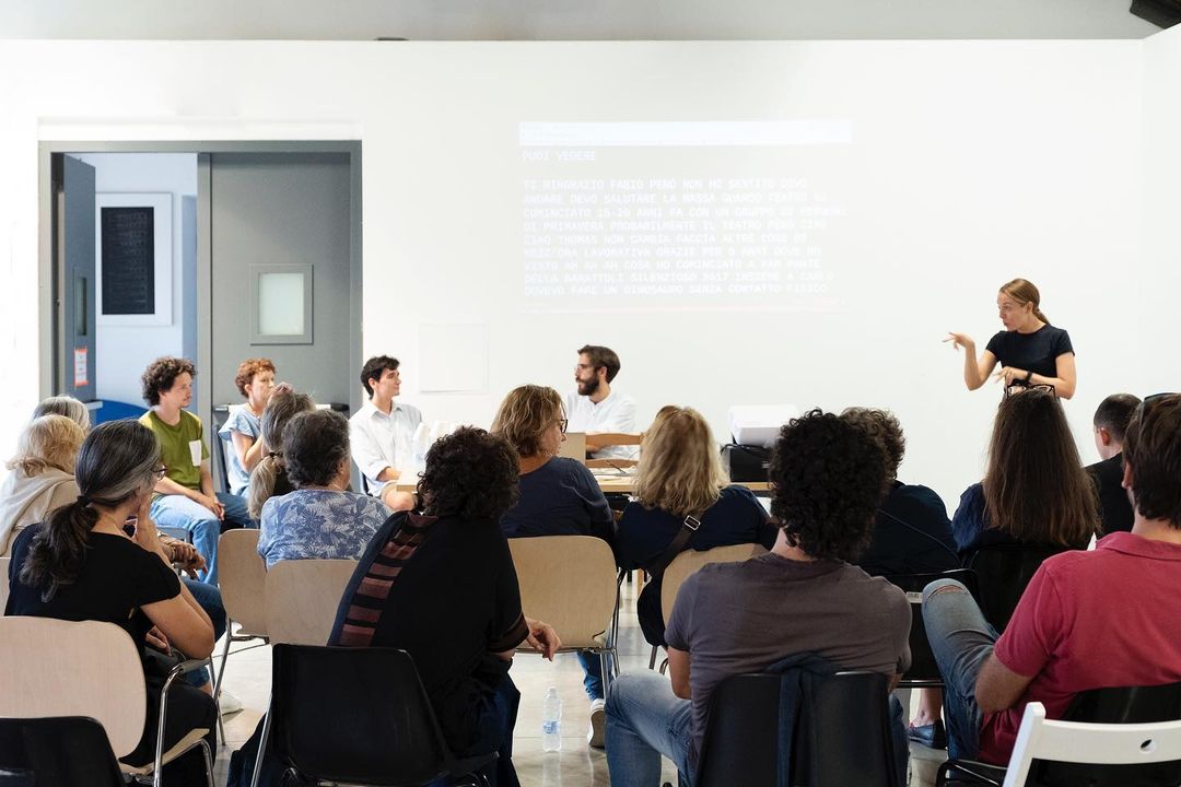 All’ interno di una sala bianca, un gruppo di persone è seduto su alcune file di sedie. Di fronte a loro alcuni relatori e relatrici intervengono. Dietro i relatori, sul muro, vengono proiettate delle slide e un’interprete traduce in LIS.