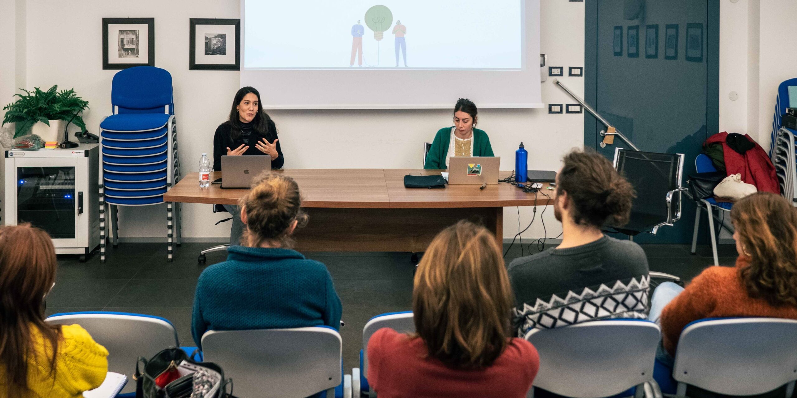 All'interno di una sala, due donne conducono un incontro sedute dietro una scrivania. Dietro di loro un'immagine proiettata. Davanti a loro sono sedute tre persone, due donne e un uomo